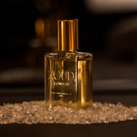 A bottle of Moresqe Elixir perfume oil with a gold cap, surrounded by scattered crystals on a dark surface.